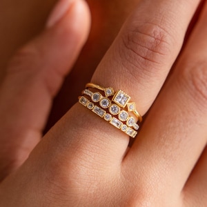May include: A hand displays three gold rings with various gemstone settings. The rings feature round, square, and baguette-cut stones. The gold bands have a textured appearance, and the gemstones sparkle against the warm lighting.