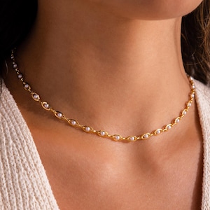 May include: A delicate gold necklace featuring small, evenly spaced pearls. The necklace is a choker style, resting closely against the neck. The gold chain has an oval link design. The pearls are white and add a touch of elegance.