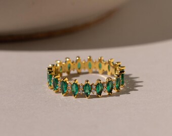 Può includere: Un anello d'oro con una fila di pietre preziose verdi a forma di marquise. L'anello è posto su una superficie bianca.