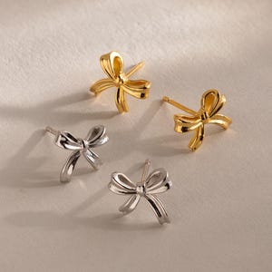 May include: Four bow-shaped stud earrings. Two are gold-colored, and two are silver-colored. The earrings are small and delicate, with a simple, elegant design. The earrings are displayed on a light-colored surface.