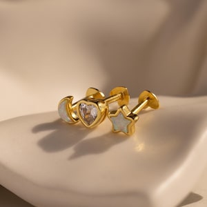 Puede incluir: Pendientes dorados con diseños de luna, corazón y estrella. Los pendientes tienen cierre de tuerca y están adornados con piedras preciosas blancas y transparentes. Se exhiben sobre una superficie blanca y curvada.