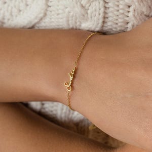 Pode incluir: Uma delicada pulseira de corrente em tom dourado com um elemento central de placa com nome escrito em letra cursiva. A pulseira consiste numa fina corrente de elos de metal polido que se prende à volta do pulso.