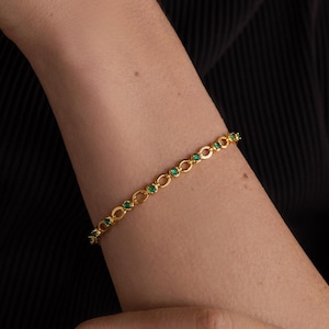以下が含まれることがあります： 小さな緑色の宝石が付いた金のチェーンブレスレット。ブレスレットは人の手首につけています。