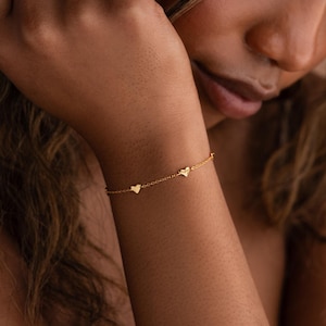 May include: A delicate gold chain bracelet featuring two small, evenly spaced heart-shaped charms. The bracelet is constructed with a fine link chain and has a polished metallic finish that reflects light, creating a subtle and refined appearance.