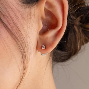 以下が含まれることがあります： 耳のクローズアップ。金色のイヤリングには、丸い透明な石が1つと、小さな透明な石がカーブを描いて並んでいます。