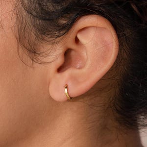 Puede incluir: Primer plano de una oreja con un pequeño pendiente de aro dorado. El pendiente tiene un diseño simple y minimalista. La piel de la persona es clara y su cabello es oscuro y rizado. El pendiente tiene aproximadamente 1,3 cm de diámetro.