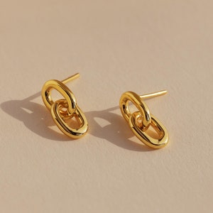 May include: A pair of gold-colored stud earrings. Each earring features two interlocked oval links. The earrings are displayed on a light beige surface, with soft shadows adding depth. The jewelry is simple and elegant.