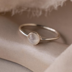 May include: A delicate silver ring featuring a round, iridescent moonstone. The ring's band is thin and polished, and the moonstone is set in a simple bezel. The ring is displayed on a neutral-toned, textured surface.
