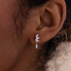 May include: A close-up of a silver earring with a floral design. The earring features multiple small, clear gemstones arranged in a cluster, with a single teardrop-shaped gem dangling below. The earring is worn on an ear.