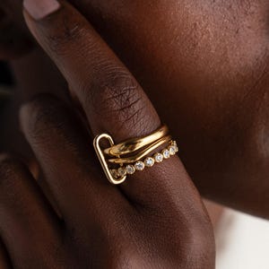May include: A close-up of a person's hand wearing three gold rings. One ring has a unique, curved design, while another features a row of small, clear stones. The rings are set against the backdrop of the person's skin, creating a contrast of textures and tones.
