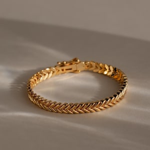 May include: A gold-toned bracelet featuring a repeating herringbone or wheat-like chain link pattern. The bracelet is circular in shape and includes a visible rectangular clasp mechanism at the top, reflecting light across its polished metallic surface.