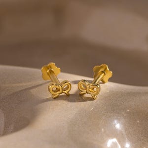 Pode incluir: Um par de brincos pequenos, dourados, em forma de laço. Os brincos são exibidos em uma superfície de tons neutros, com um fundo macio e desfocado. Os laços têm desenhos detalhados de nós e laços.