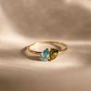 May include: A gold ring featuring two pear-shaped gemstones set side by side. One gemstone is a light blue color, and the other is a deep olive green. The band is thin and polished, creating a delicate look with the two contrasting stones.