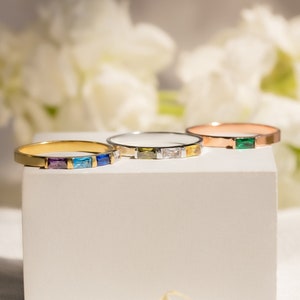 May include: Four gold rings with colorful gemstones on a white display stand. The rings are stacked on top of each other, with the largest ring at the bottom and the smallest ring at the top. The gemstones are blue, green, yellow, and clear.