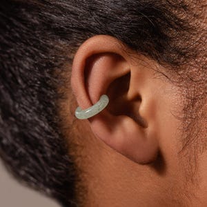 May include: A smooth, pale green circular ear cuff made of a solid material. The cuff features a polished finish and a simple, continuous band design that rests securely along the outer edge of the ear cartilage, providing a subtle accessory.