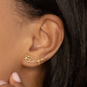 Dainty gold stud name earrings sit it in the first piercing of the ear. The charm earring has the name 'Raegan' spelled in a minimalist script font.