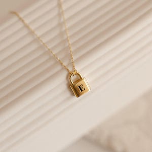 May include: A gold-toned necklace featuring a delicate chain and a small, rectangular padlock pendant. The front of the padlock is engraved with the capital letter E in a serif font. The metal has a polished finish and a warm yellow hue.