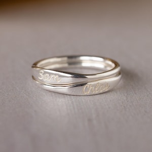 May include: A pair of silver rings, one atop the other, with a smooth, polished finish. The top ring is engraved with the name "Sam", and the bottom ring has the name "Chloe". The rings are set against a neutral background.