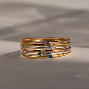 May include: A stack of five thin gold, silver, and rose gold rings with small colourful gemstones. The gemstones are a light blue, green, pink, and purple.