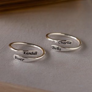 Könnte beinhalten: Zwei silberfarbene Wickelringe. Ein Ring ist mit "Kendall" und "River" graviert, der andere mit "Charlie" und "Bella". Die Ringe liegen auf einer hellgrauen Oberfläche, mit einem verschwommenen goldenen Objekt im Hintergrund.