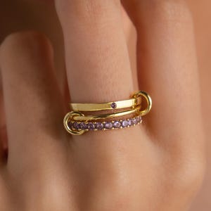 Könnte beinhalten: Drei goldene Ringe sind auf einem Finger gestapelt. Der obere Ring hat einen kleinen lila Stein, während der mittlere Ring mit einer Reihe kleiner lila Steine verziert ist. Die Ringe sind mit kleinen goldenen Schlaufen verbunden.