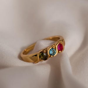 Może przedstawiać: Złoty pierścionek z trzema kolorowymi kamieniami szlachetnymi. Pierścionek ma gładką, zaokrągloną obrączkę i zawiera zielony, niebieski i czerwony kamień.