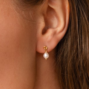 May include: A close-up of a gold-colored earring with a small, white pearl. The earring is attached to a person's ear. The earring has a small loop design at the top.