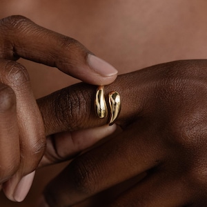 Könnte beinhalten: Ein goldfarbener Ring mit einem einzigartigen, organischen Design. Der Ring umschließt den Finger mit zwei geschwungenen, tropfenförmigen Elementen. Der Ring wird am Finger einer Person gezeigt, mit dem Hautton im Hintergrund.