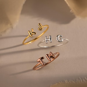 May include: Three delicate rings, each featuring a different set of initials. One ring is gold-toned with "K J", another is silver-toned with "H G", and the third is rose gold-toned with "T N". The rings are displayed on a neutral background.
