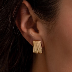 Puede incluir: Primer plano de un pendiente rectangular dorado con detalles de barras verticales. El pendiente se lleva en la oreja de una persona con cabello oscuro y tez clara. El pendiente tiene un diseño moderno y geométrico.