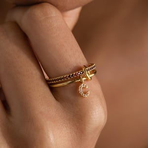 以下が含まれることがあります： ゴールドリングセット。小さな赤い石が並んだバンドと、結び目のデザインのバンドがあります。小さなゴールドのチャームは、文字「C」の形で、小さなクリアストーンがちりばめられています。リングは指にあります。