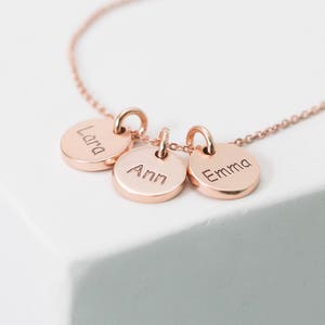 May include: A rose gold necklace with three round charms, each engraved with a name: Lara, Ann, and Emma.