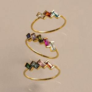 May include: Three gold rings with colorful gemstones. Each ring features a row of rectangular gemstones in different colors, including blue, green, pink, and red.