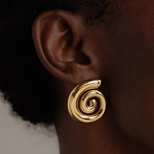 Puede incluir: Primer plano de un pendiente en espiral dorado. El pendiente es de metal pulido con una superficie lisa. El diseño es una forma de espiral audaz y escultural, con una parte superior redondeada y un interior arremolinado.