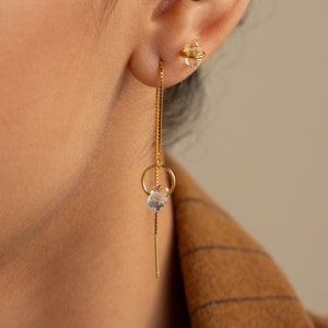 Puede incluir: Un pendiente de cadena dorada con un dije de cristal transparente y un pequeño aro de oro. El pendiente cuelga del lóbulo de la oreja de una persona con cabello oscuro.