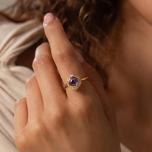 May include: A gold ring featuring a central round purple gemstone set within a hexagonal frame. The hexagonal setting is accented with small, clear, sparkling stones along its edges, mounted on a thin, polished gold band.