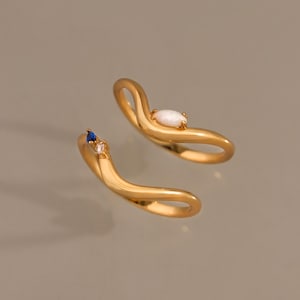 May include: A gold open-style ring featuring a wavy, fluid band design. One end is set with a single oval-shaped white opal, while the other end features a small, deep blue gemstone paired with a tiny, clear, rectangular-cut stone.