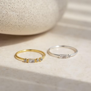 May include: Two thin rings, one in gold-tone metal and one in silver-tone metal. The gold ring features a central baguette-cut clear stone flanked by two small round clear stones. The silver ring features a central baguette-cut clear stone.