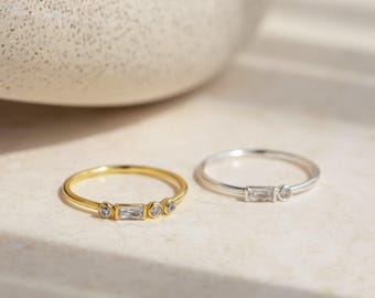 May include: Two thin rings, one in gold-tone metal and one in silver-tone metal. The gold ring features a central baguette-cut clear stone flanked by two small round clear stones. The silver ring features a central baguette-cut clear stone.