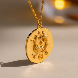 May include: Gold-colored pendant necklace featuring a round, textured pendant with an engraved design. The pendant is suspended from a delicate chain. The design on the pendant includes two mythical creatures and small gemstones. The necklace is displayed on a light-colored surface.