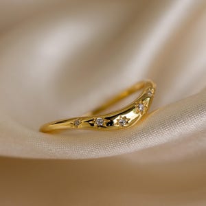 May include: A curved gold ring with star-shaped and round clear gemstones. The ring is set against a soft, cream-colored background, highlighting its delicate design. The ring's band curves gently, creating an elegant and modern look.