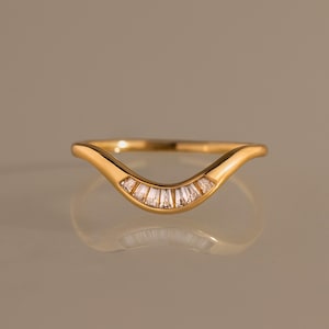 May include: A gold curved ring featuring a series of small, clear baguette-cut stones set along the center dip of the band. The polished metal band has a smooth, rounded finish that follows a gentle V-shape contour, highlighting the stones.