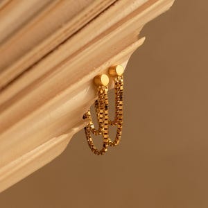 May include: A pair of gold-toned earrings featuring a small circular stud connected to a delicate box chain that loops downward to form a dangling hoop shape. The earrings have a polished metallic finish and a simple, clean design structure.