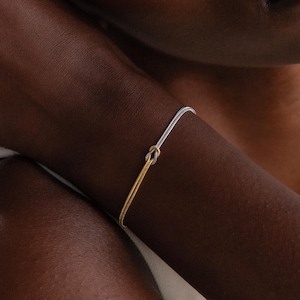 May include: A two-tone bracelet with a knot design. The bracelet features a silver band that transitions into a gold band. The jewelry is worn on the wrist. The bracelet is a delicate piece of jewellery.