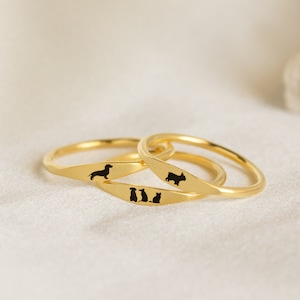 Three dainty gold rings each with an engraved pet design in black ink. The middle ring has a combination of three different pets, both dogs and cats, engrave on it which the other two have one dog engraved on each.