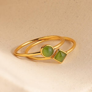 May include: Two gold rings with green gemstone accents. One ring has a round stone, the other has a square stone.