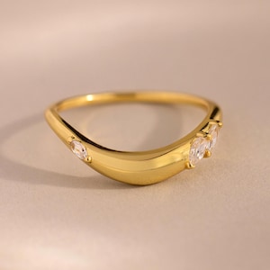 May include: A gold-colored ring with a curved design. The ring features three clear, marquise-cut gemstones. The band has a smooth, polished finish. The ring is set against a soft, neutral background.