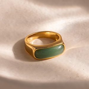 May include: A gold-colored ring with a rectangular, light green stone set in the center. The ring has a smooth, polished finish and is displayed on a white surface. The ring's design is simple and elegant, suitable for everyday wear.