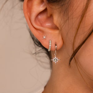 May include: A silver hoop earring with a small star charm dangling from it. The hoop is set with small green gemstones. The earring is worn in a person's ear.