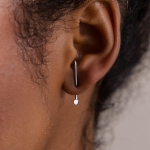 Puede incluir: Primer plano de una oreja con un pendiente plateado. El pendiente tiene una barra recta que atraviesa la oreja y un pequeño disco redondo que cuelga debajo del lóbulo. El tono de piel es medio.